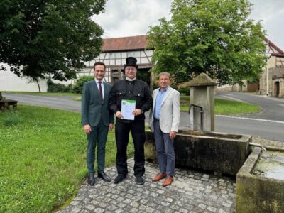 Landrat Florian Töpper (links) und Stadtlauringens erster Bürgermeister Friedel Heckenlauer (rechts) gratulierten Kaminkehrermeister Gerald Raab zum erneuten Erhalt der Urkunde des Klima- und Umweltpakts Bayern. Das Bild entstand beim Dorfbrunnen in Altenmünster vor dem Hintergrund des neu gestalteten Pilgerhofs. Foto: Andreas Lösch/Landratsamt Schweinfurt